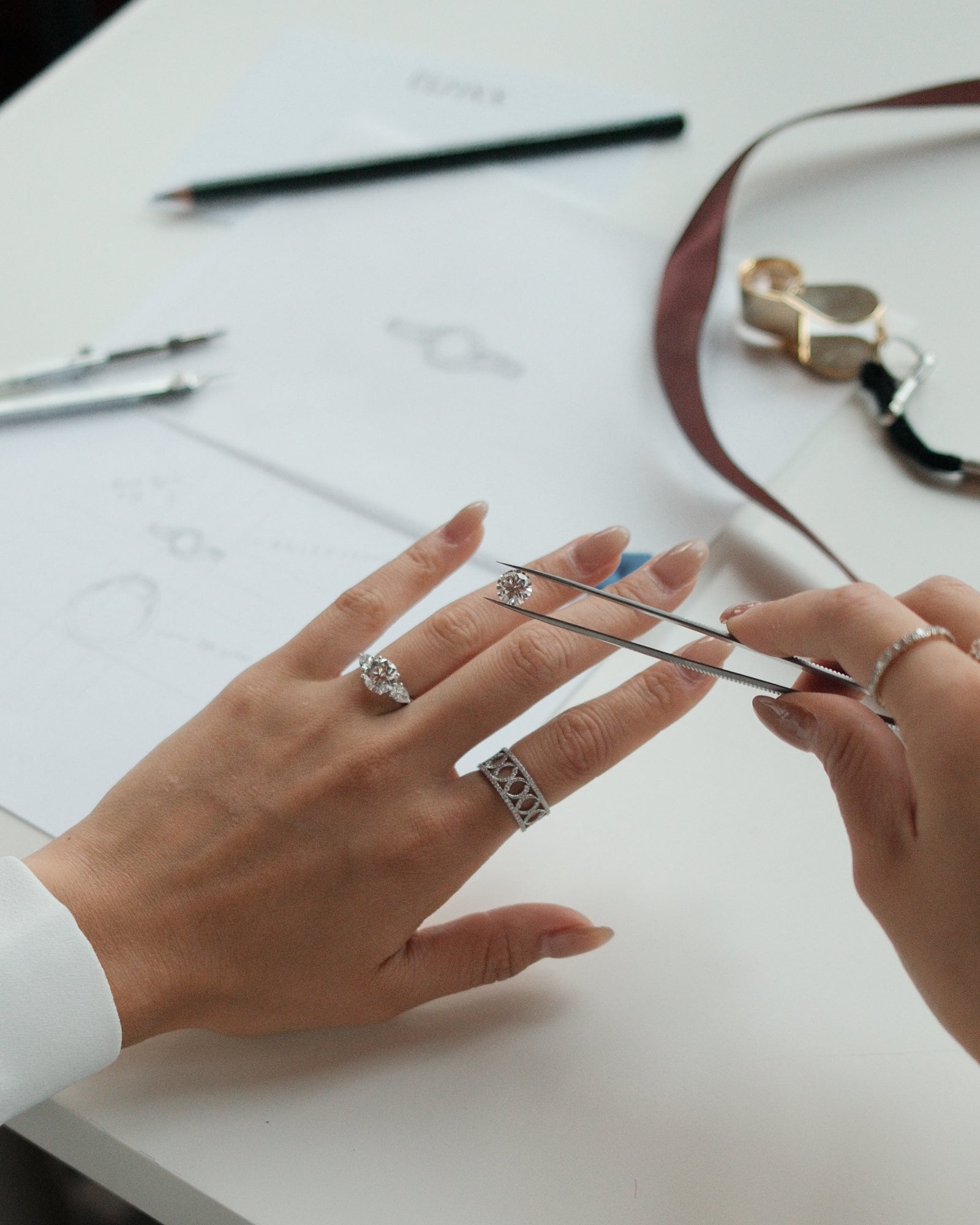 Person wearing multiple rings on a white surface with jewelry and design tools.