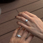 Two hands shown wearing diamond engagement rings with 18K white gold open gap stacker bands, set against a warm brown wooden plank background.