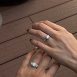 Two hands shown wearing diamond engagement rings with 18K white gold open gap stacker bands, set against a warm brown wooden plank background.