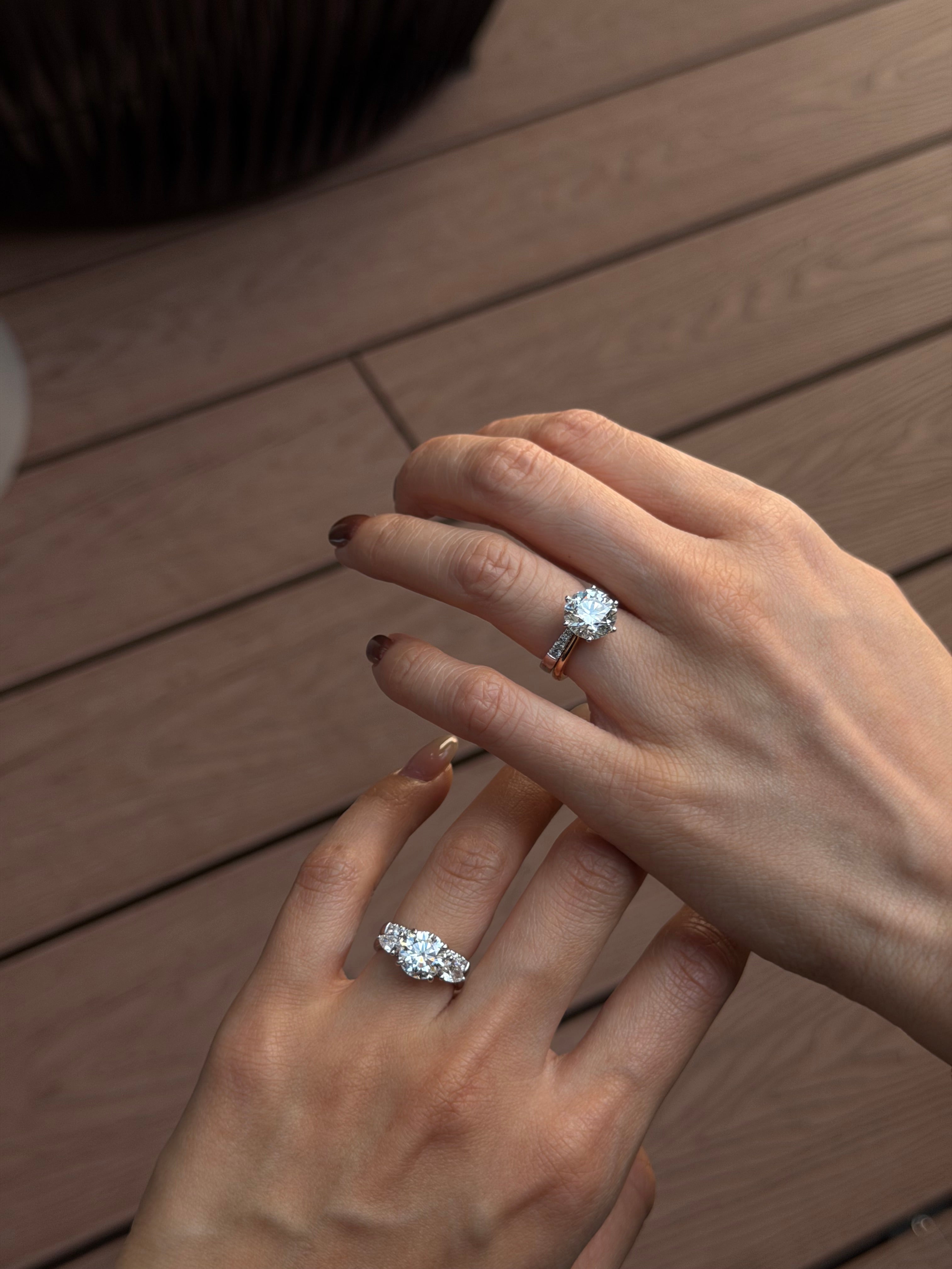 Two hands shown wearing diamond engagement rings with 18K white gold open gap stacker bands, set against a warm brown wooden plank background.