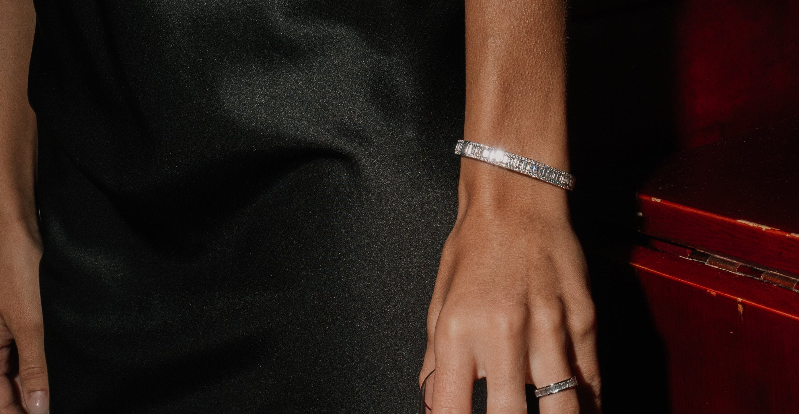 Person holding a glass of red wine with a black silk dress, tinxx baguette lab grown diamond bangle and ring, against a red background