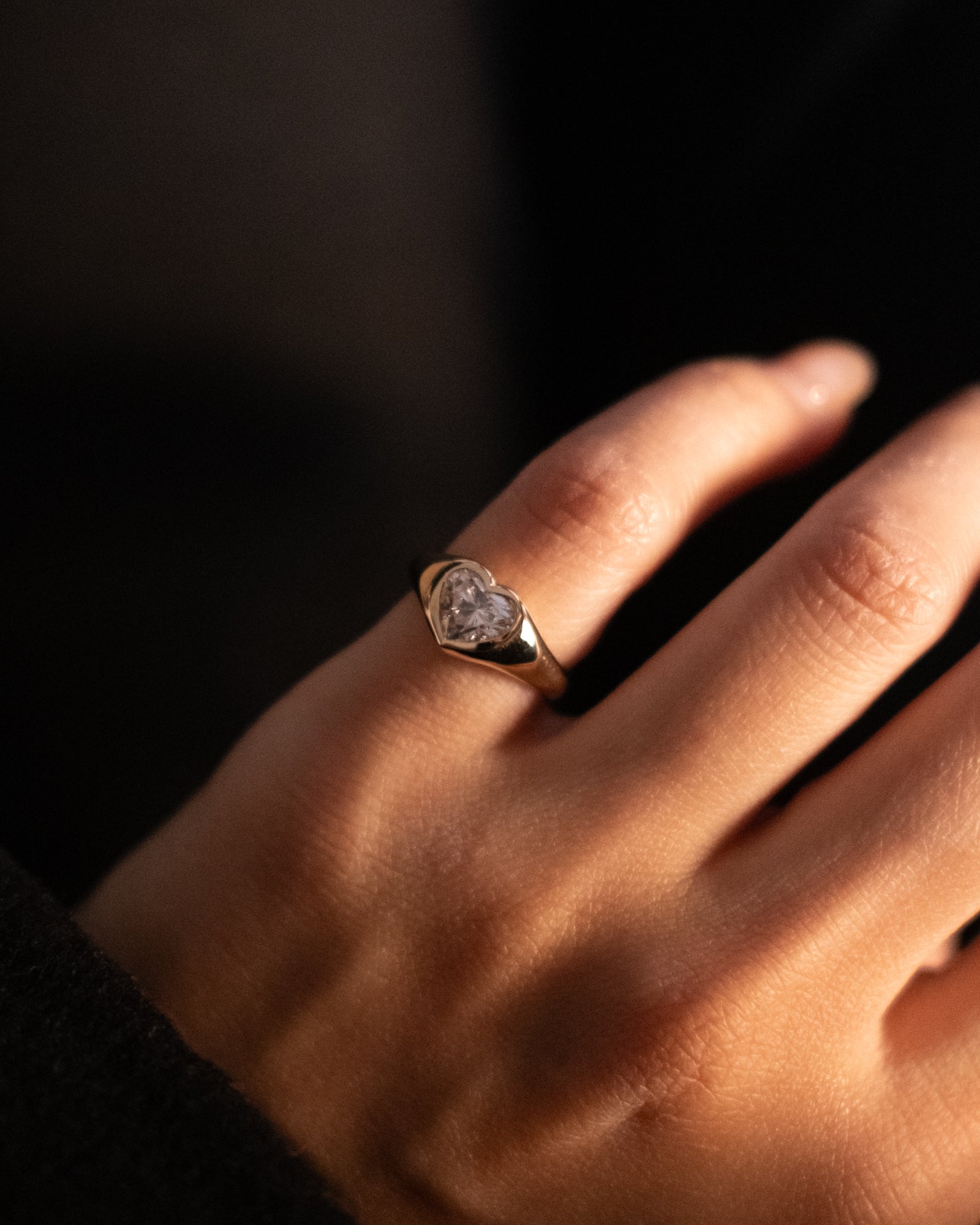 Heart shaped diamond signet ring in 18K gold on a pinky finger with a blurred background