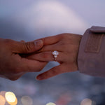 Two hands reaching towards each other with a focus on one hand wearing a tinxx jewelry engagement ring, blurred lights in the background.