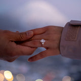 Two hands reaching towards each other with a focus on one hand wearing a tinxx jewelry engagement ring, blurred lights in the background.