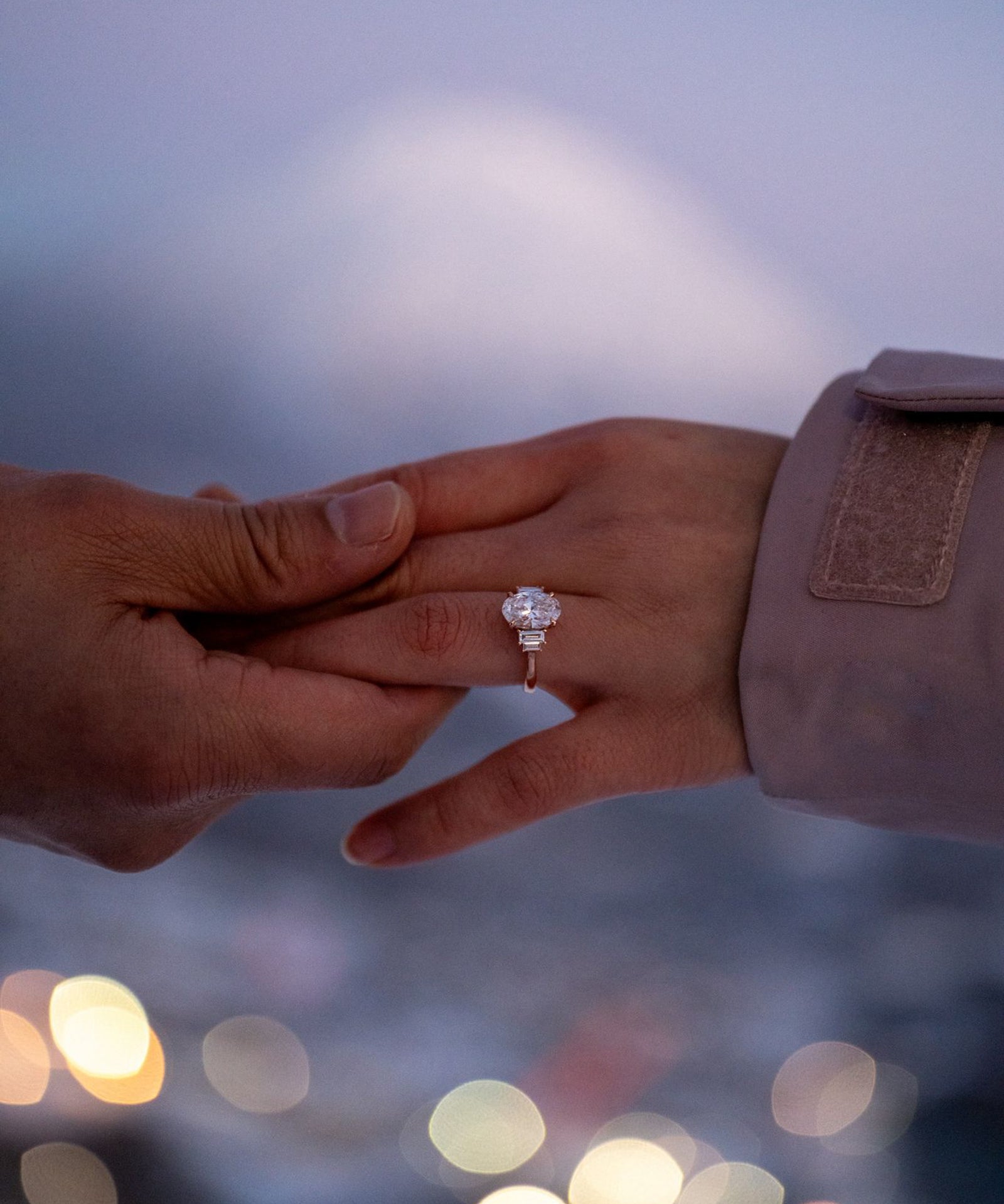 Two hands reaching towards each other with a focus on one hand wearing a tinxx jewelry oval engagement ring, blurred lights in the background.