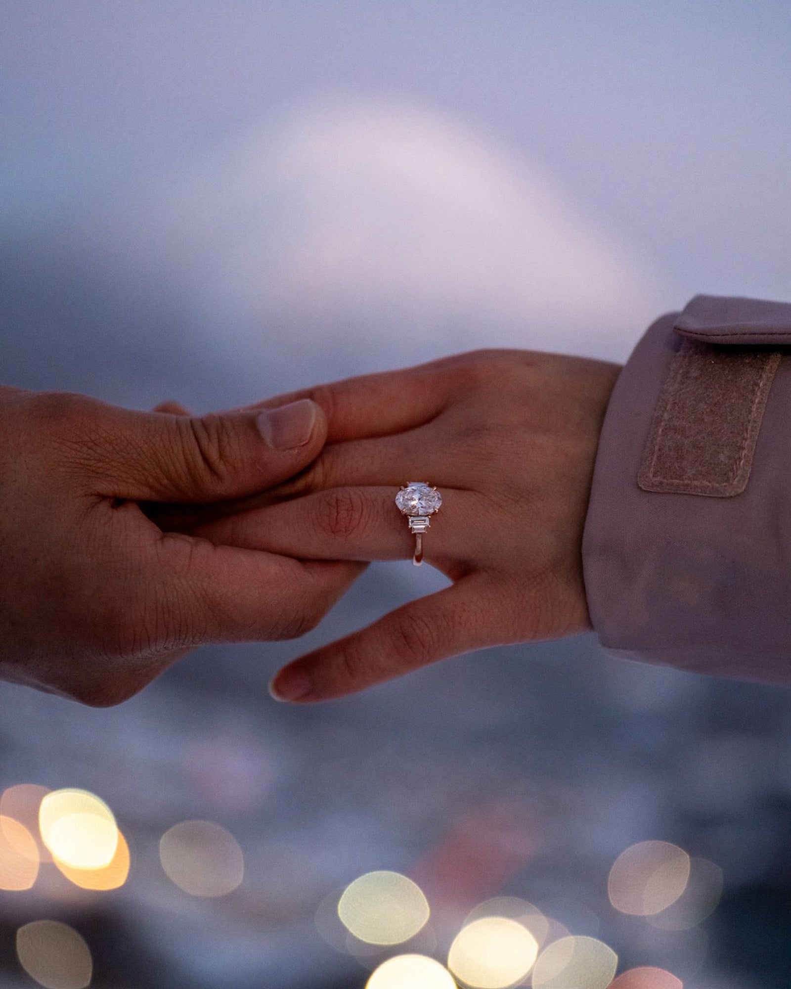 Two hands reaching towards each other with a focus on one hand wearing a tinxx jewelry engagement ring, blurred lights in the background.
