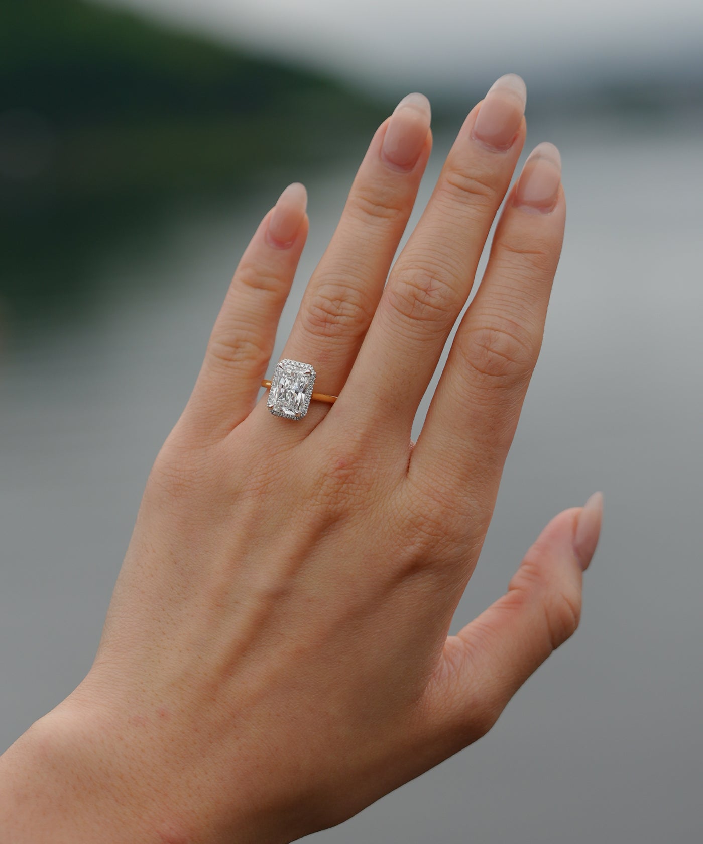 Hand wearing a tinxx lab grown radiant cut diamond halo ring with a blurred natural background