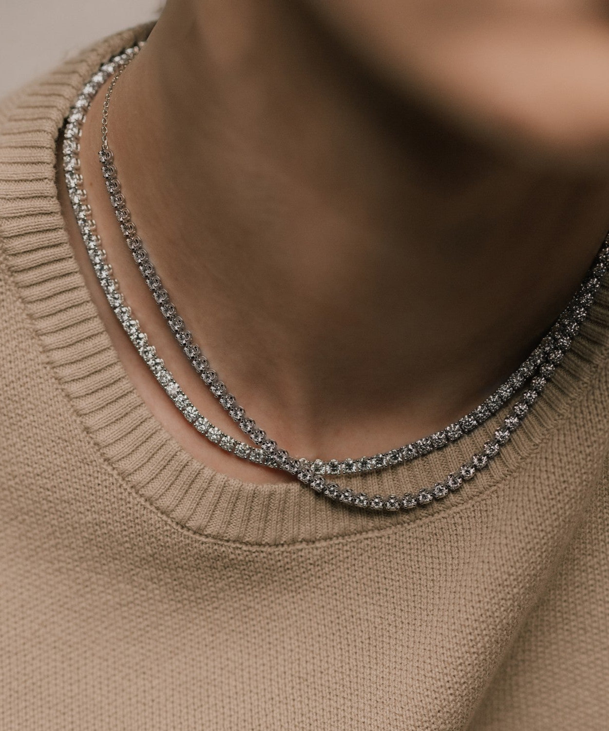 Two round cut lab grown diamond tennis necklaces layered, worn by a woman in a beige soft knit shirt, photographed in soft natural lighting from tinxx.
