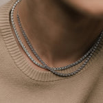 Two round cut lab grown diamond tennis necklaces layered, worn by a woman in a beige soft knit shirt, photographed in soft natural lighting from tinxx.