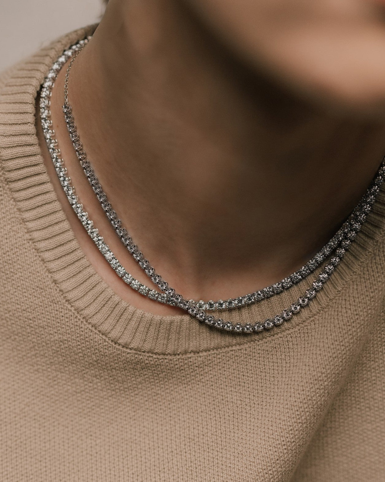 Two round cut lab grown diamond tennis necklaces layered, worn by a woman in a beige soft knit shirt, photographed in soft natural lighting from tinxx.