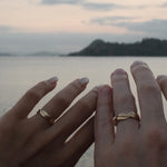 Two hands resting together with matching twist 18K yellow gold rings on the fingers, set against a soft-focus background of water and a distant island at dusk.