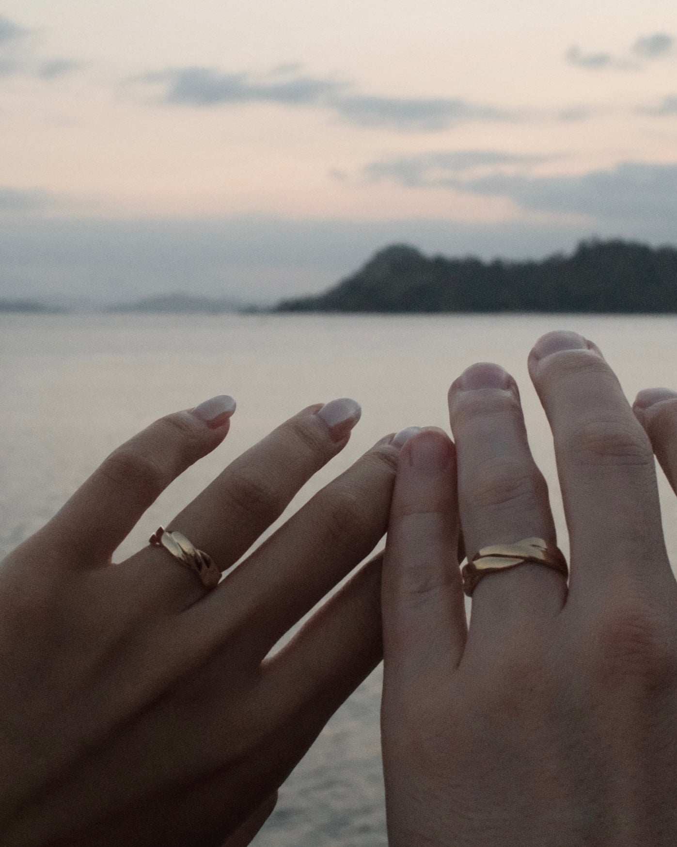 Two hands resting together with matching twist 18K yellow gold rings on the fingers, set against a soft-focus background of water and a distant island at dusk.