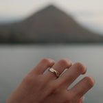 A close-up of a hand wearing a delicate twist band 18K yellow gold ring, with a blurred mountain and water in the background.