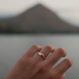A close-up of a hand wearing a delicate twist band 18K yellow gold ring, with a blurred mountain and water in the background.