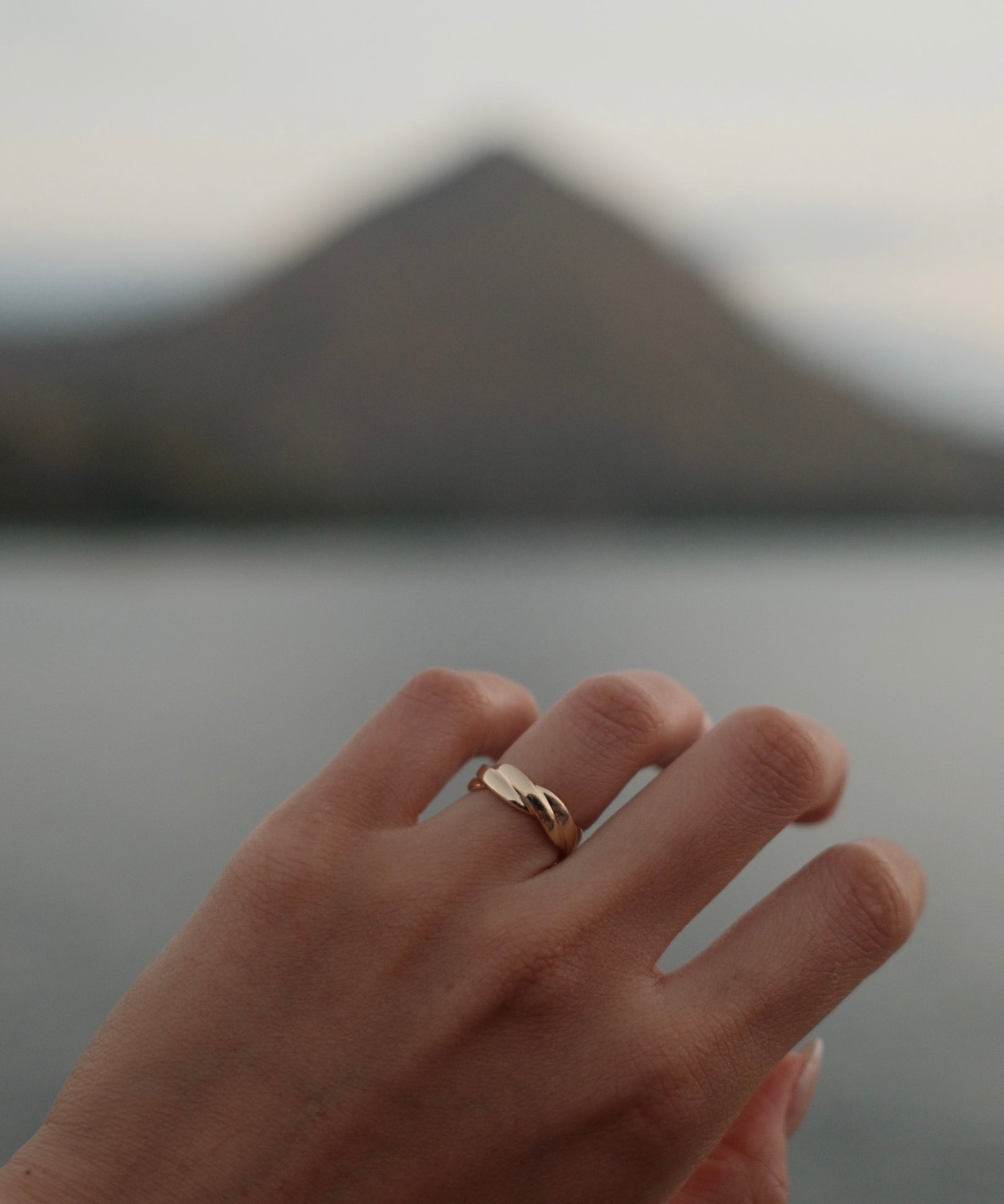 A close-up of a hand wearing a delicate twist band 18K yellow gold ring, with a blurred mountain and water in the background.