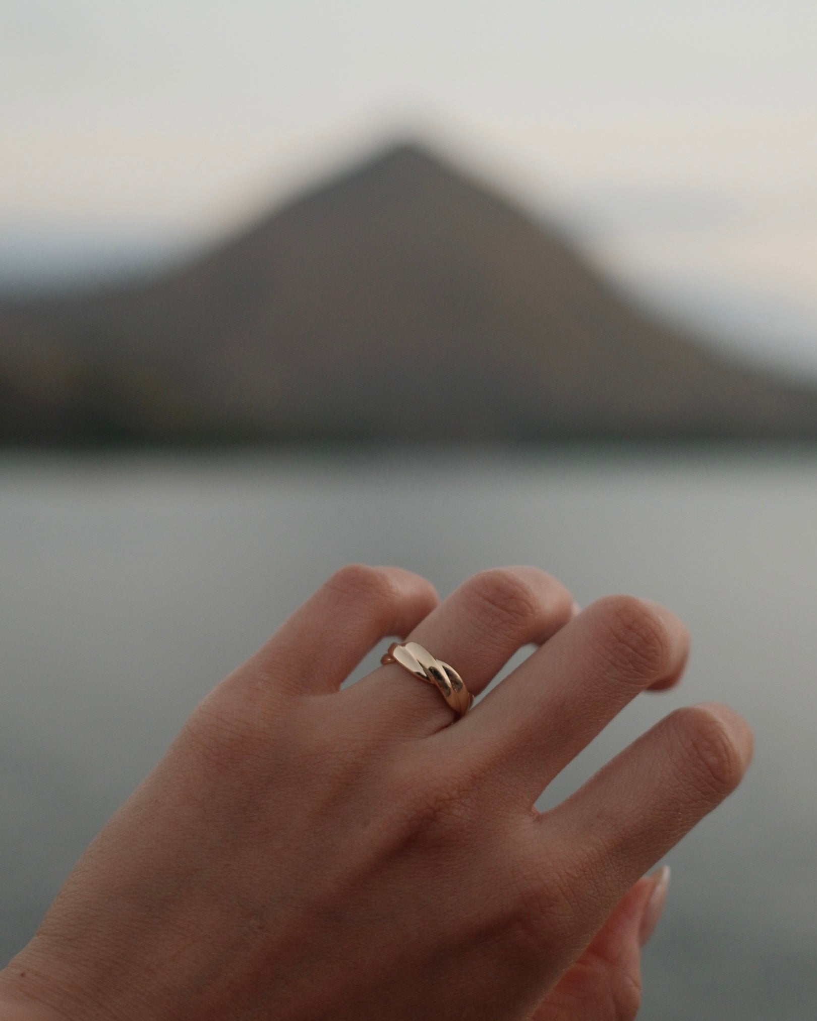 A close-up of a hand wearing a delicate twist band 18K yellow gold ring, with a blurred mountain and water in the background.
